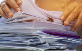 The image displays a close-up of hands sorting through a large stack of papers and documents, suggesting an extensive filing or archiving task. The papers are of various thicknesses and sizes with some having visible tabs and labels. The person handling the documents appears to be in the middle of organizing or searching for specific information within the pile. This visual context is evocative of the meticulous process involved in document conversion services, where physical documents are prepared for digitization and conversion into electronic formats, a critical step in modern information management and digital archiving.