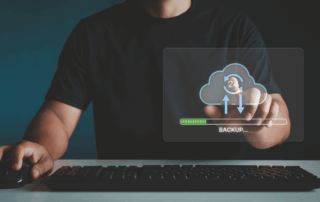 Man sits behind a keyboard and mouse with a conceptual icon that he is pointing to or clicking. The icon has a cloud and 24 signifying that backups are done daily. This is a Data Backup and Recovery conceptual image.